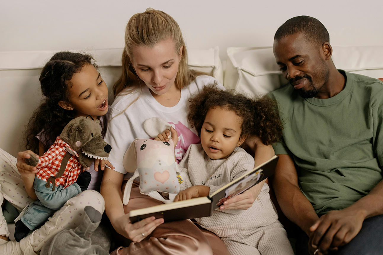 Two adults and two children sit together on a sofa reading a book
